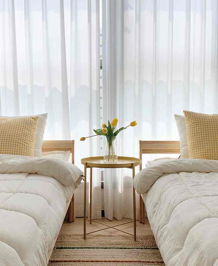 White and natural wood tone bedroom featuring yellow check pillows and a nightstand with yellow tulip vase creating a cozy space