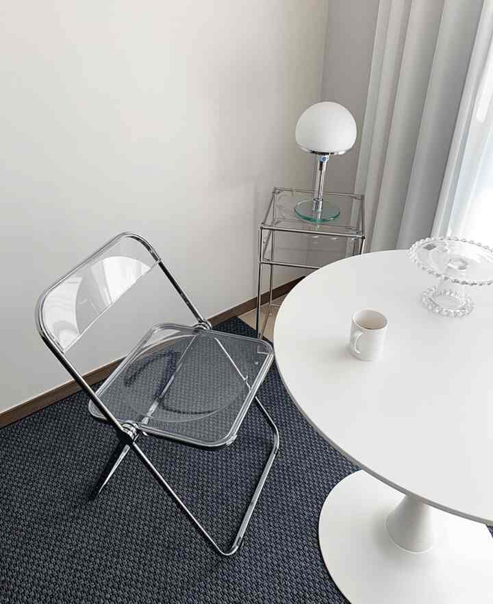 White-toned living room featuring a transparent folding chair, round dining table, and Bauhaus-style side table with table lamp in a clean setting