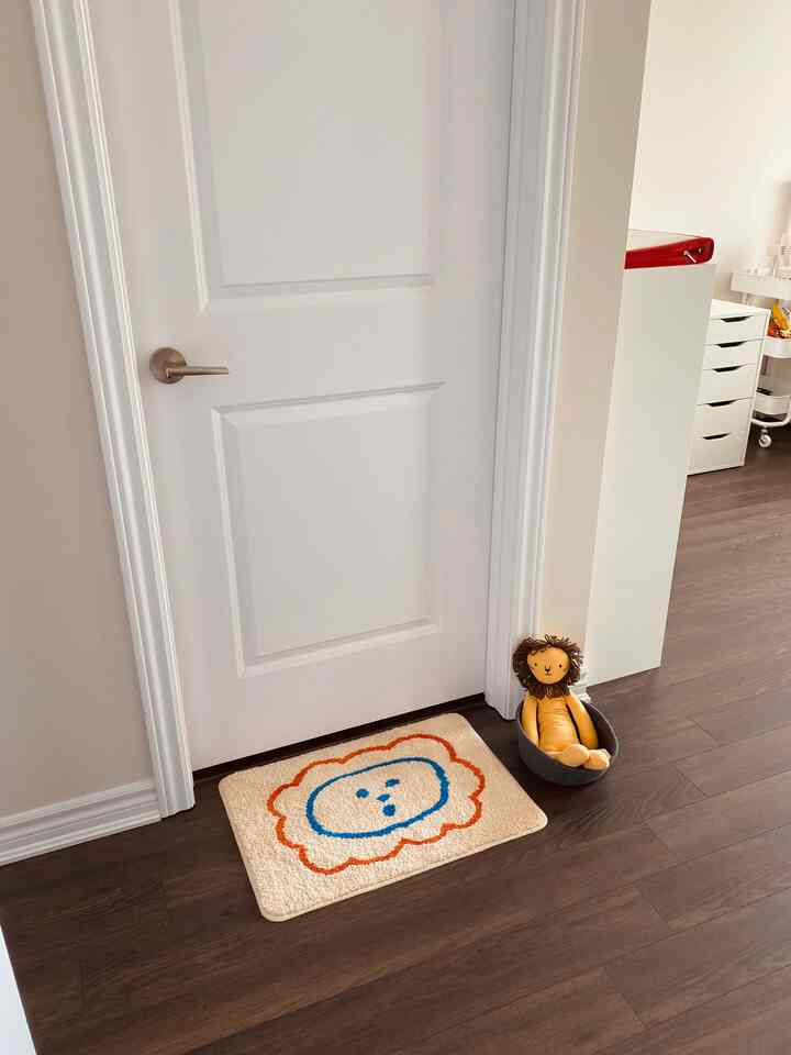 白い扉とナチュラルウッドの床材、かわいいバスマットとライオンのぬいぐるみが配置されたバスルームの入り口