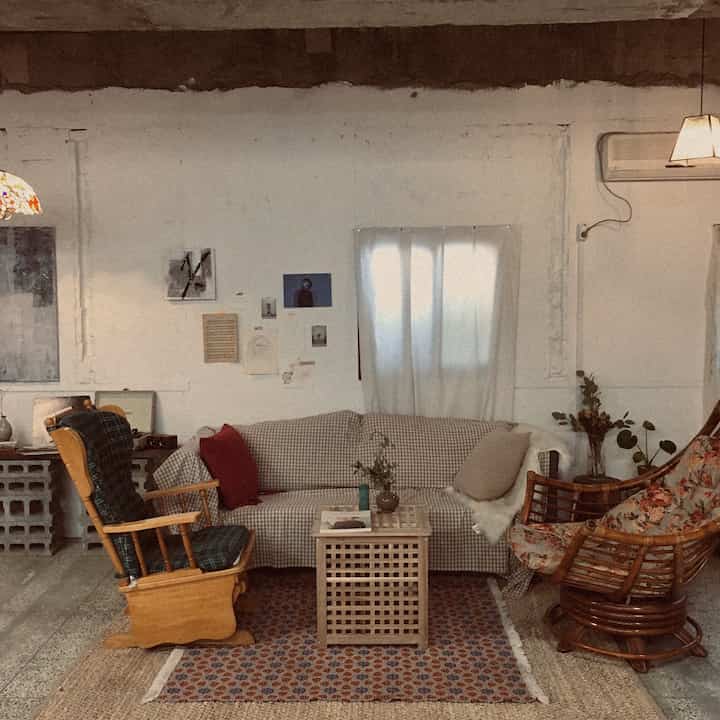 Beige and natural wood tone living room featuring a checkered sofa, rattan chair, and wooden storage table creating a cozy, vintage studio space