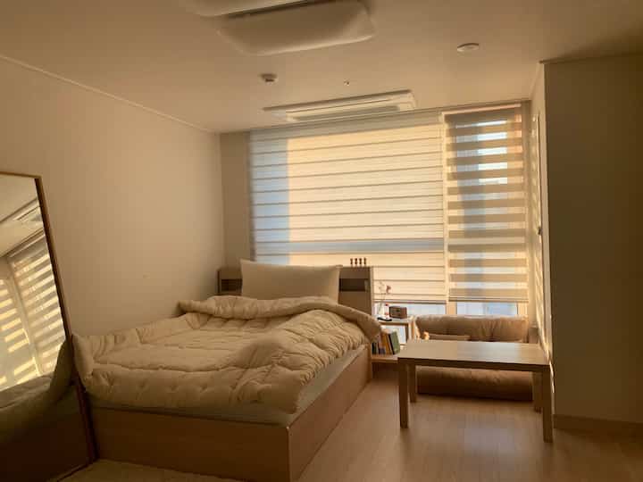 温かみのあるベージュと木目調のワンルーム寝室、ベッドとナイトテーブル、全身鏡が配置された落ち着いた空間