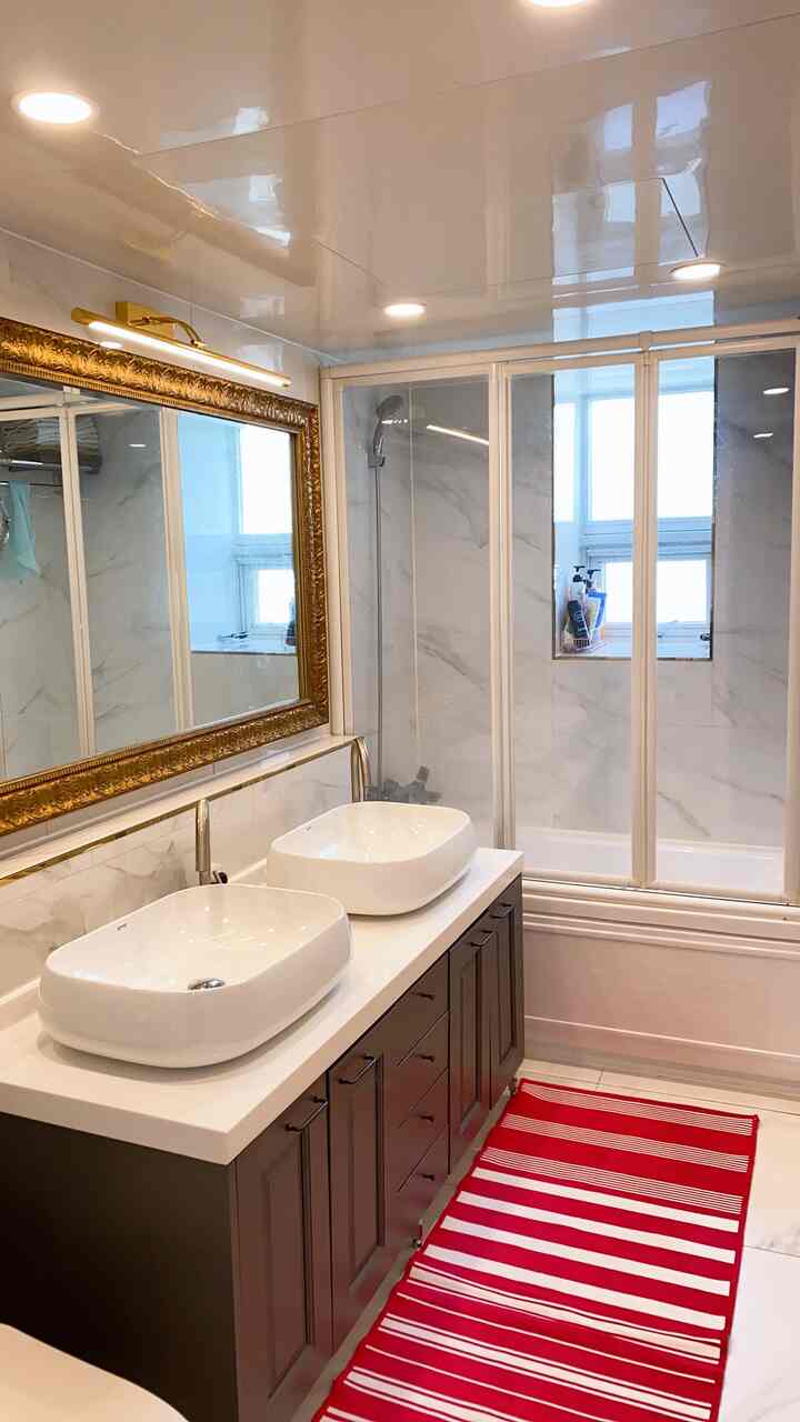 White and brown tone bathroom featuring large washbasins, a gold-framed mirror, shower booth, and a red striped bath mat in a clean setting