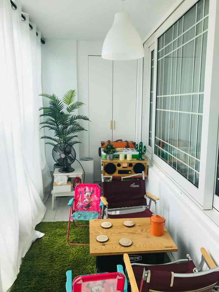 Bright natural-tone balcony featuring kids' chairs, green IKEA rug, pendant light, and wooden table creating cozy home cafe ambiance