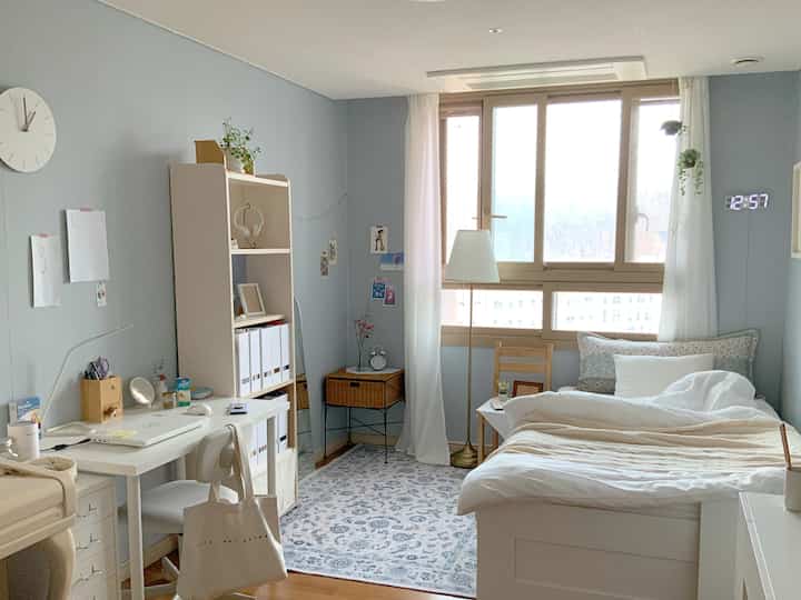 ブルーグレーの色調が特徴的な一人暮らしの寝室、机とベッド、自然な木製の窓枠が配置された清潔感のある雰囲気のインテリア