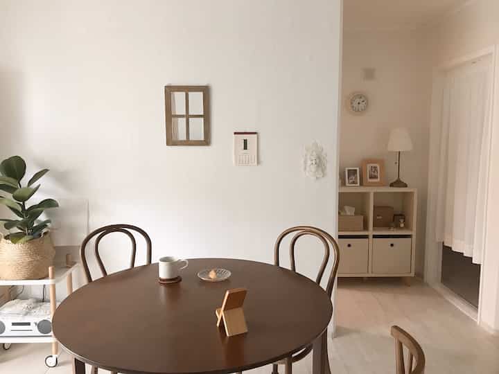 White and brown toned living dining area featuring solid wood round table, chair set, and rattan plant basket creating cozy natural vibe