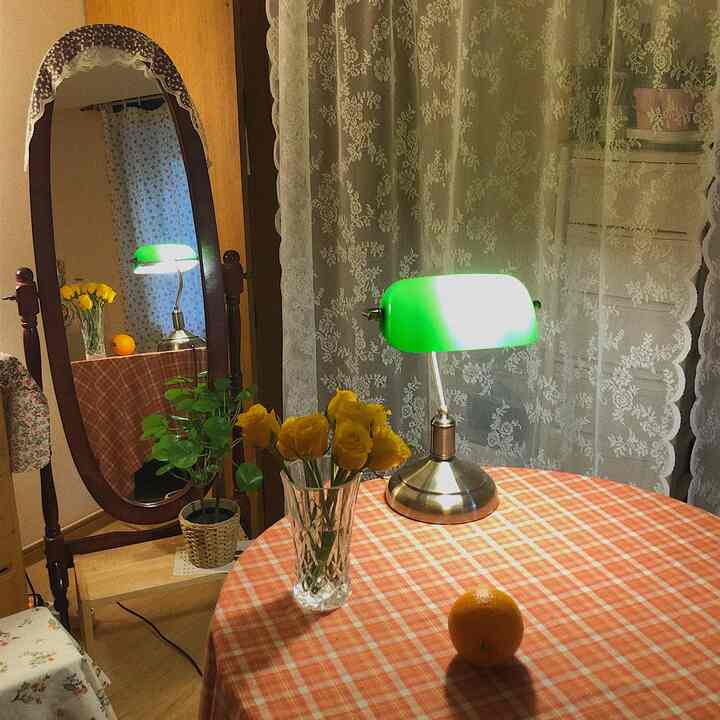 Cozy vintage-style bedroom featuring a round table with yellow roses in a vase, green desk lamp, and lace curtains in the background