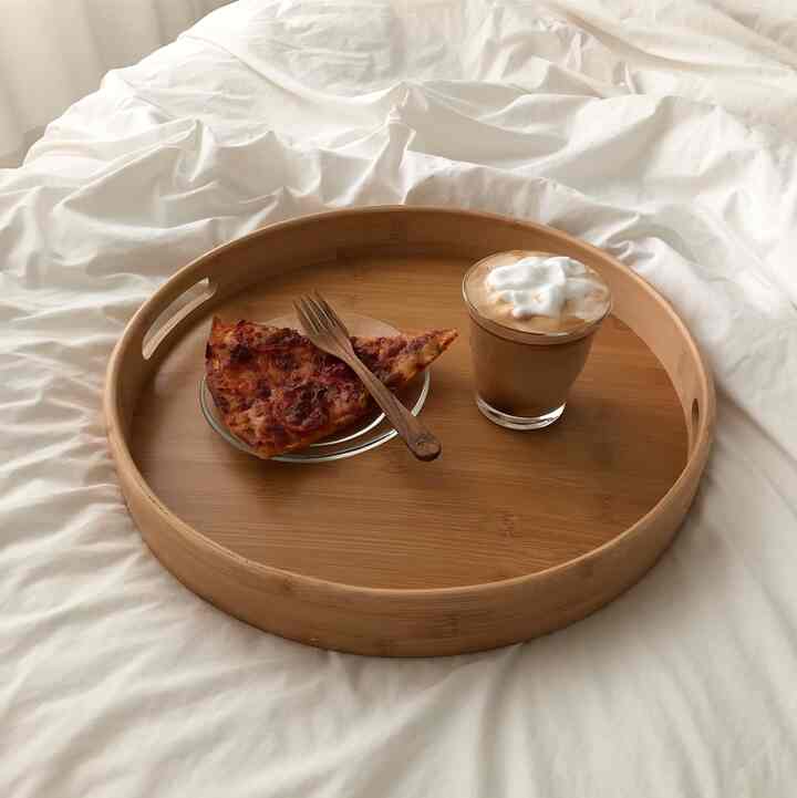 Cozy bedroom scene with white bedding featuring a wood-tone round tray holding home cafe drink and pizza slice