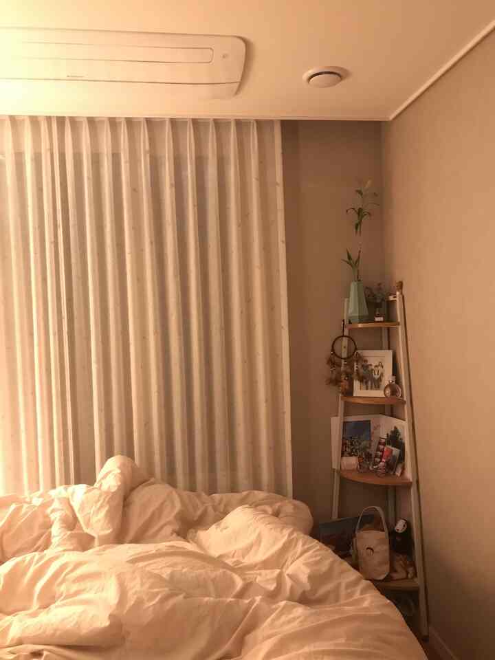 A cozy bedroom with beige walls and white bedding, custom curtains on the window, and a corner shelf enhancing space utilization