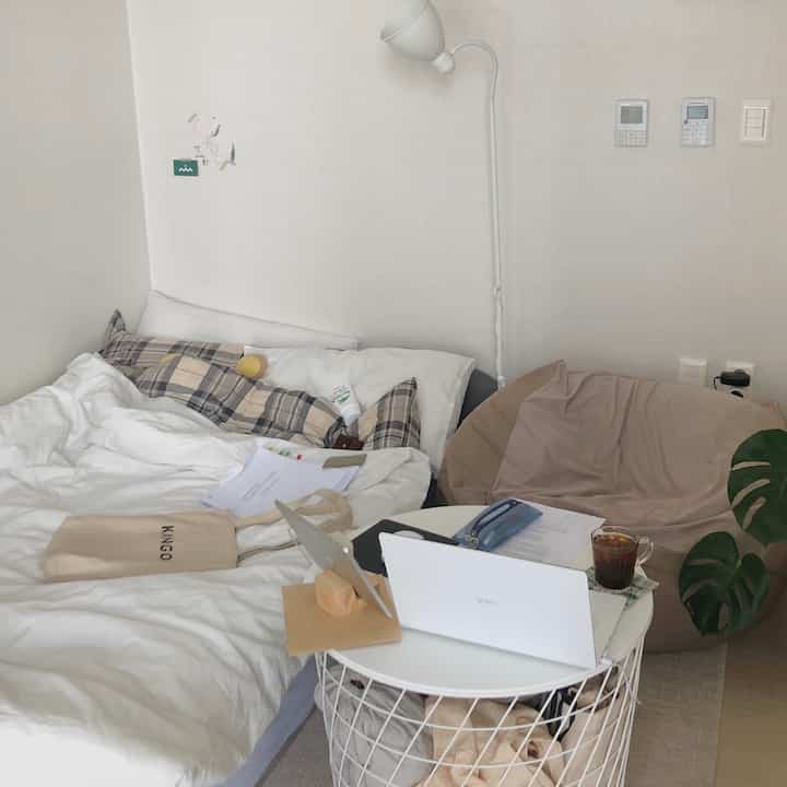 White and beige toned studio bedroom with bed and round desk featuring a laptop, creating a cozy remote work space