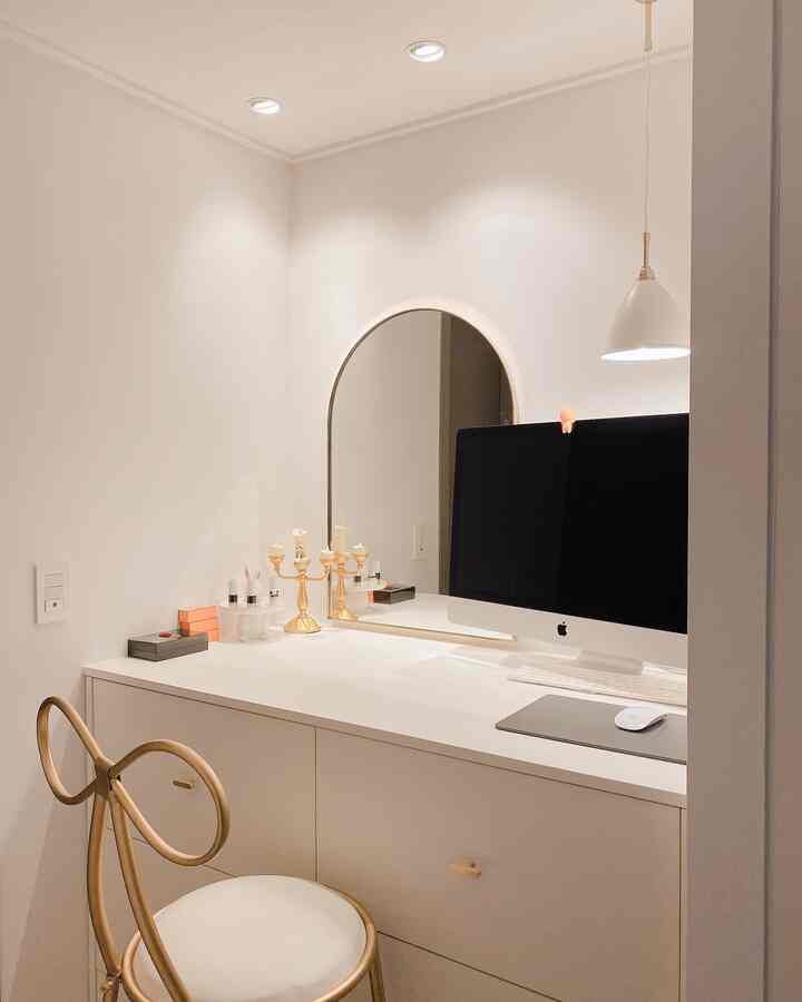 白とゴールドのトーンのモダンなホームオフィス、化粧台とアーチ型の鏡、ゴールドのスツールが配置されたミニマルなインテリア
