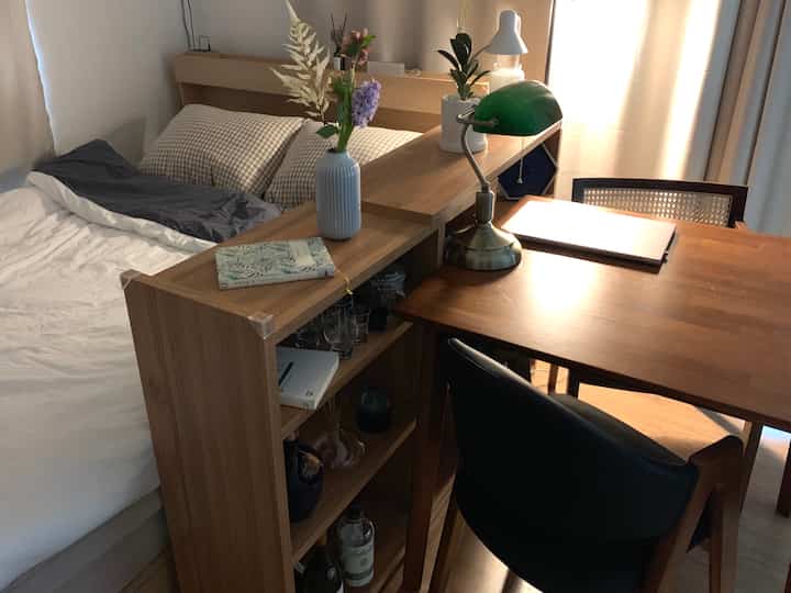 ナチュラルな木目調のワンルーム、間仕切り家具とダイニングテーブル、ラタンチェア、本、デスクランプが配置された雰囲気のある作業と休憩の空間