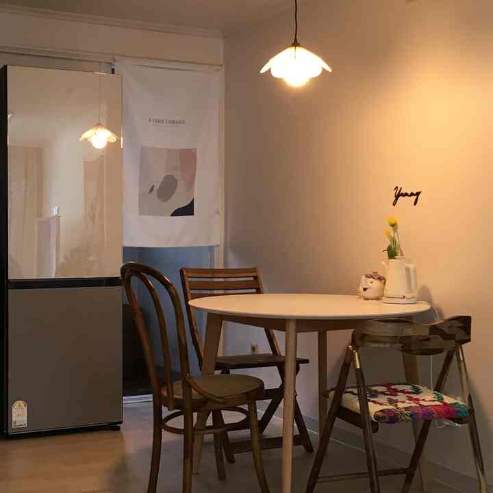 White-walled dining room featuring natural wood chairs and a round table for two, accented by warm pendant light and fabric poster near kitchen