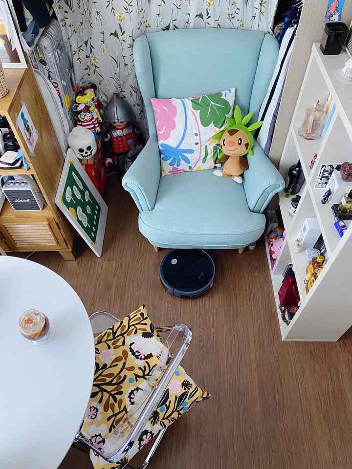 白と木目調の色調のワンルームリビング、ライトティール色のアームチェアと花柄クッション、ロボット掃除機が配置された雰囲気のある空間