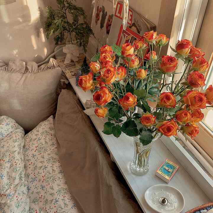 A warm beige and brown toned bedroom window area featuring a vase of vivid orange-yellow roses and cozy cushions creating a relaxing ambiance
