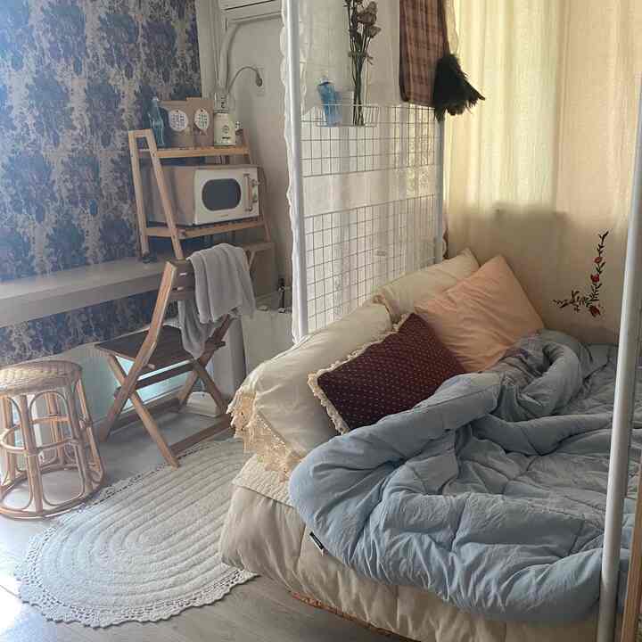 Light beige natural-toned studio apartment featuring folding chair, rug, curtains, and bedding with cozy atmosphere