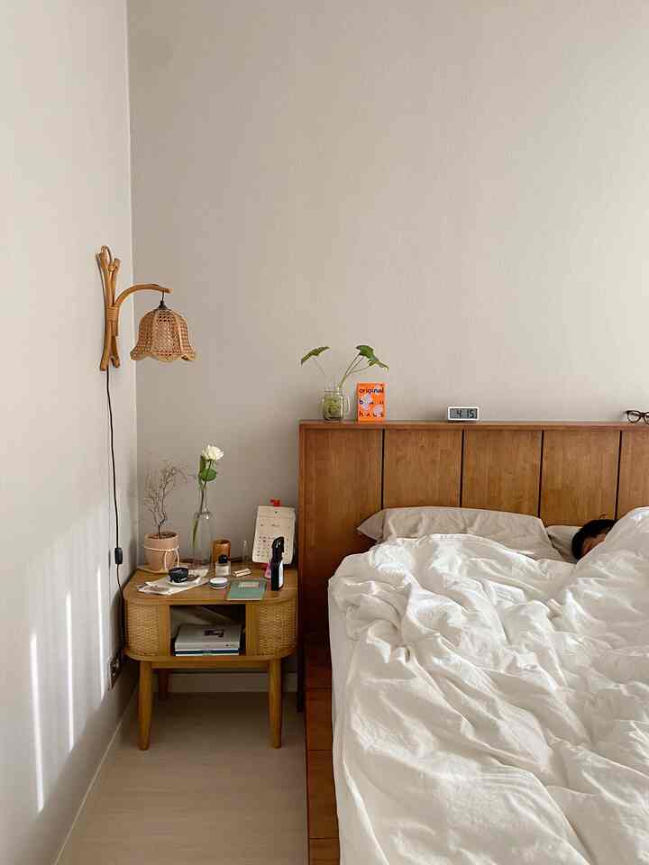 ナチュラルなブラウンの木製ベッドとホワイトの寝具が配置された寝室、ラタンのナイトテーブルと壁掛け照明が温かみのある雰囲気を演出するインテリア