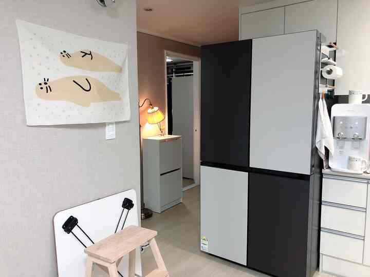 ベージュトーンの壁と明るい木製フロアが調和する玄関とキッチンの空間に、可愛らしいファブリックポスターとサムスンビスポーク冷蔵庫が配置された印象的なインテリア