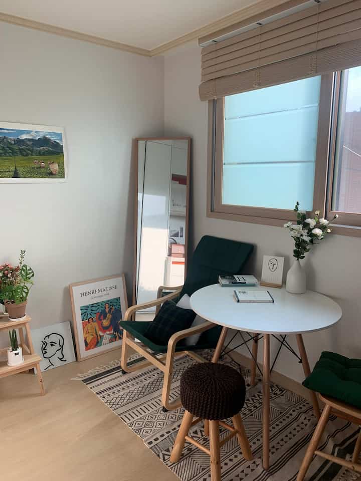 A natural-style small studio room with light brown flooring, wood tone furniture, green cushions, and plants arranged neatly