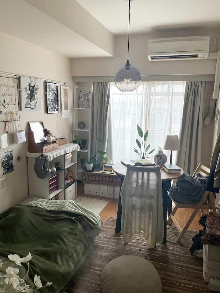 ナチュラルな色調のコンパクトなワンルームリビング兼寝室。丸いダイニングテーブル、ペンダントライト、カーテン、植物が配置された落ち着いた空間