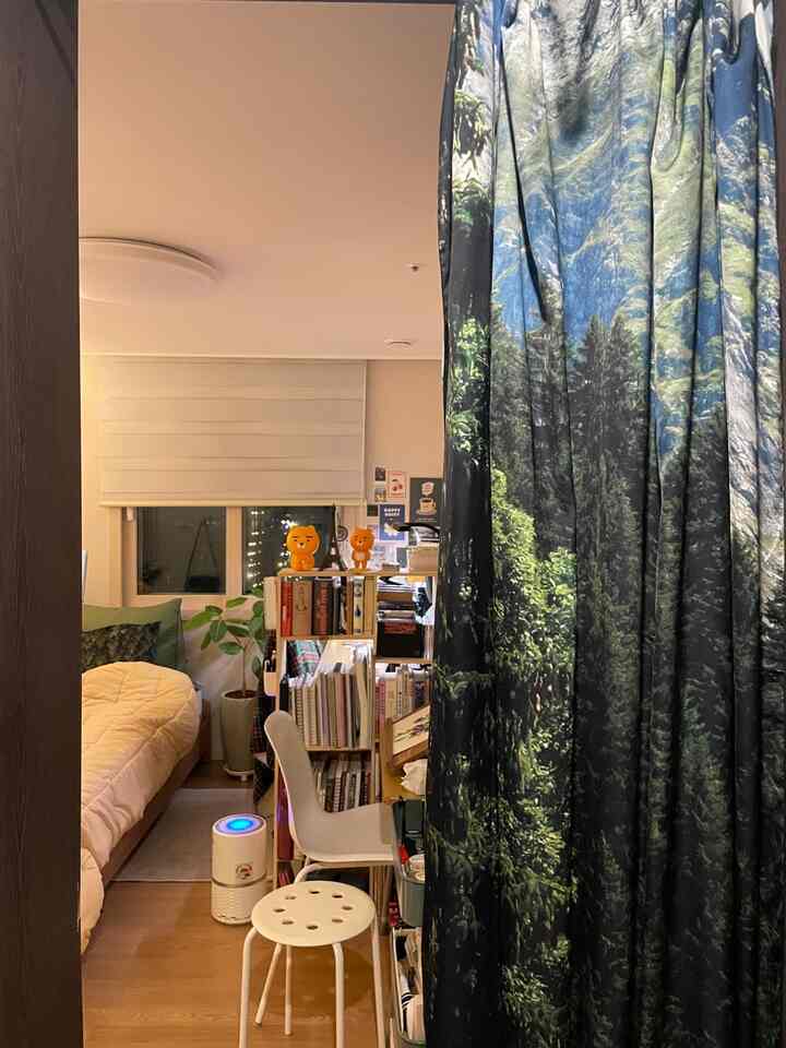 Cozy small bedroom with white walls, wooden flooring, forest-image curtain, desk, and stool arranged neatly