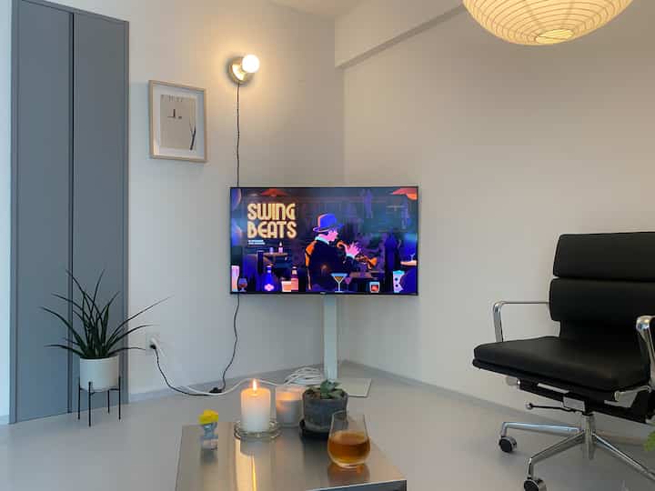 白を基調としたコンパクトなリビング、黒のオフィスチェアとブラケットライトが配置されたミニマルな空間