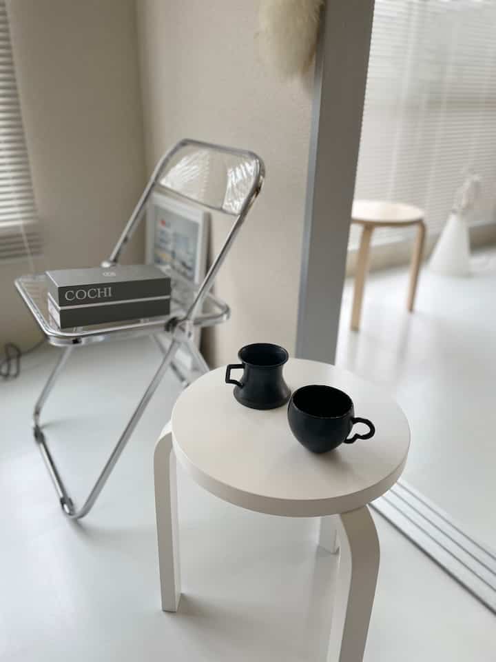 白を基調とした小さな室内空間、透明な折りたたみ椅子と白い丸いスツールの上に2つの黒いマグカップが置かれたミニマルでカフェ風の雰囲気のあるインテリア
