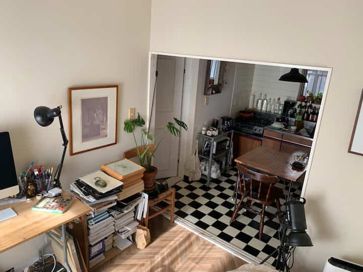 白壁と木目調の家具が調和するリビングとキッチンがつながる空間、デスクやダイニングテーブル、植物が配置された温かみのあるインテリア