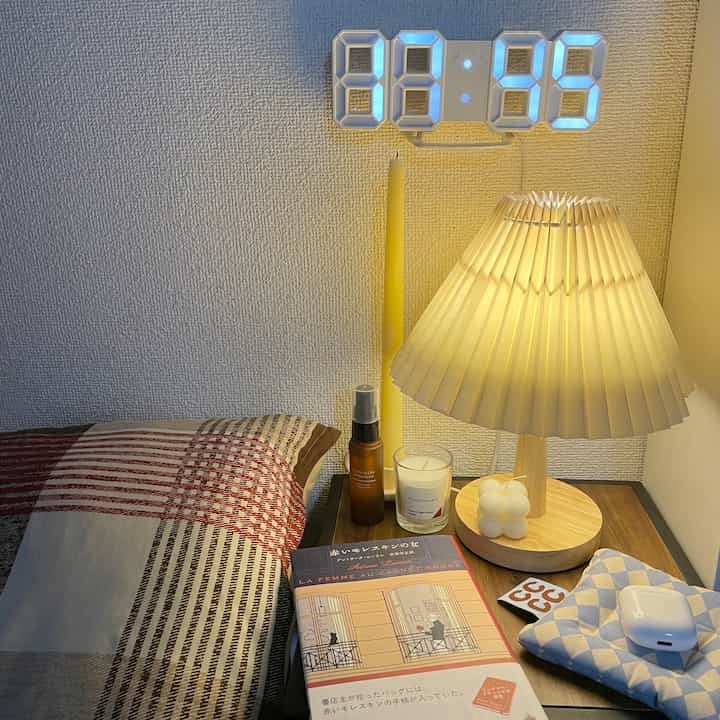 白い壁と木目調のテーブルが配置された寝室、テーブルランプとキャンドル、読書用の小説が置かれたシンプルで落ち着いた雰囲気のインテリア