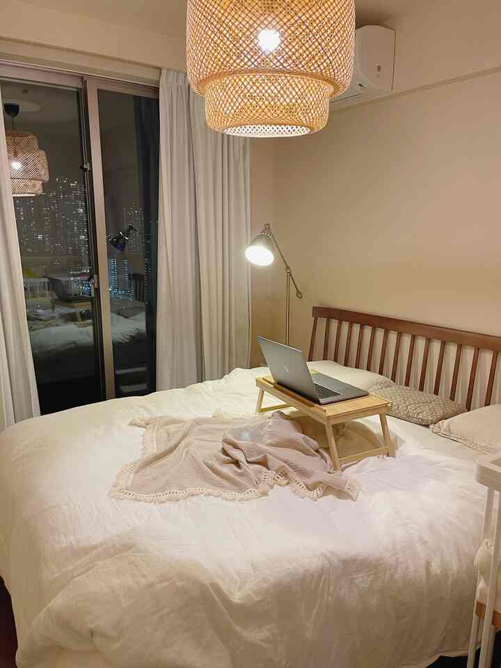Cozy bedroom with natural wood furniture and beige walls, featuring white bedding and a laptop on a wooden tray