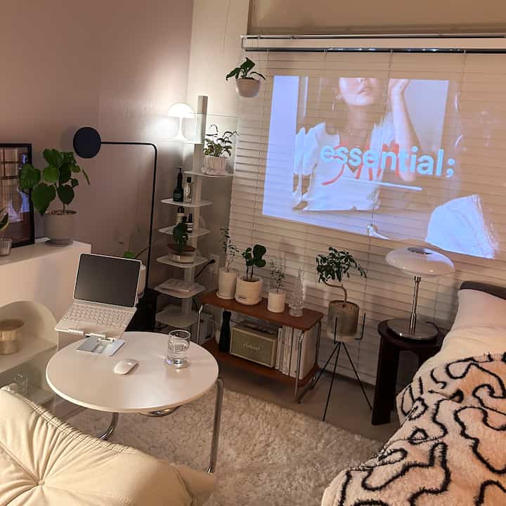 暖かみのあるベージュとホワイトの色調のリビング、ソファと小さな丸テーブルにノートパソコンが置かれ、多くの植物と照明が居心地の良い雰囲気を演出しているインテリア