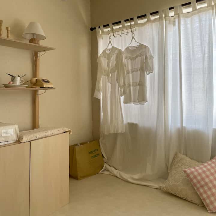 温かみのあるベージュと白のトーンのワンルームリビング、ナチュラルな木製収納とカーテン、ハンガーにかかった衣服が配置された雰囲気のある空間