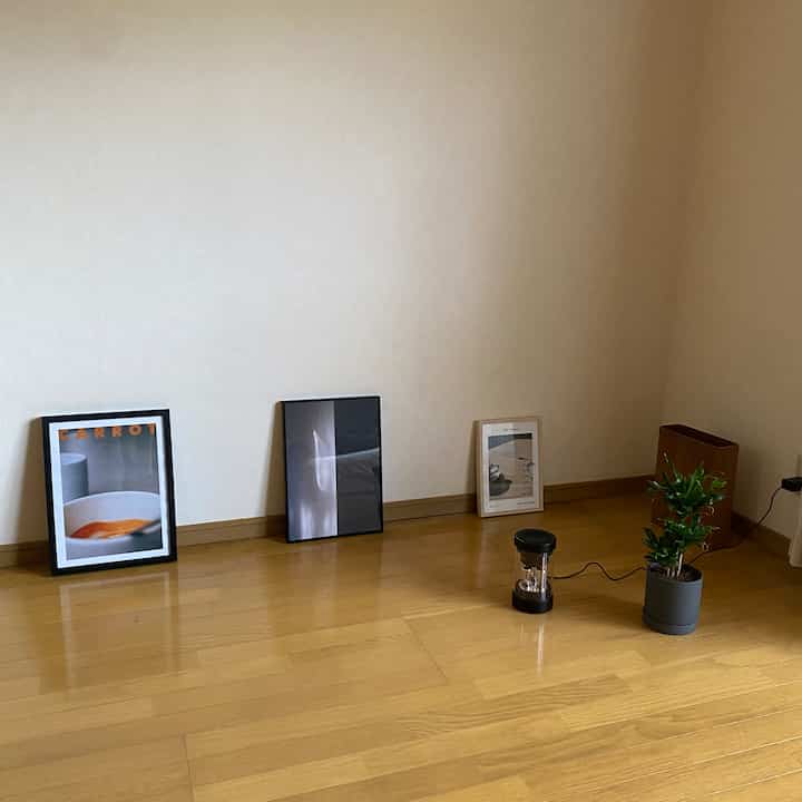 木目調の床とベージュの壁が融合したコンパクトな部屋、床に立てかけられた3つのポスターと小さな観葉植物が配置されたミニマルな空間