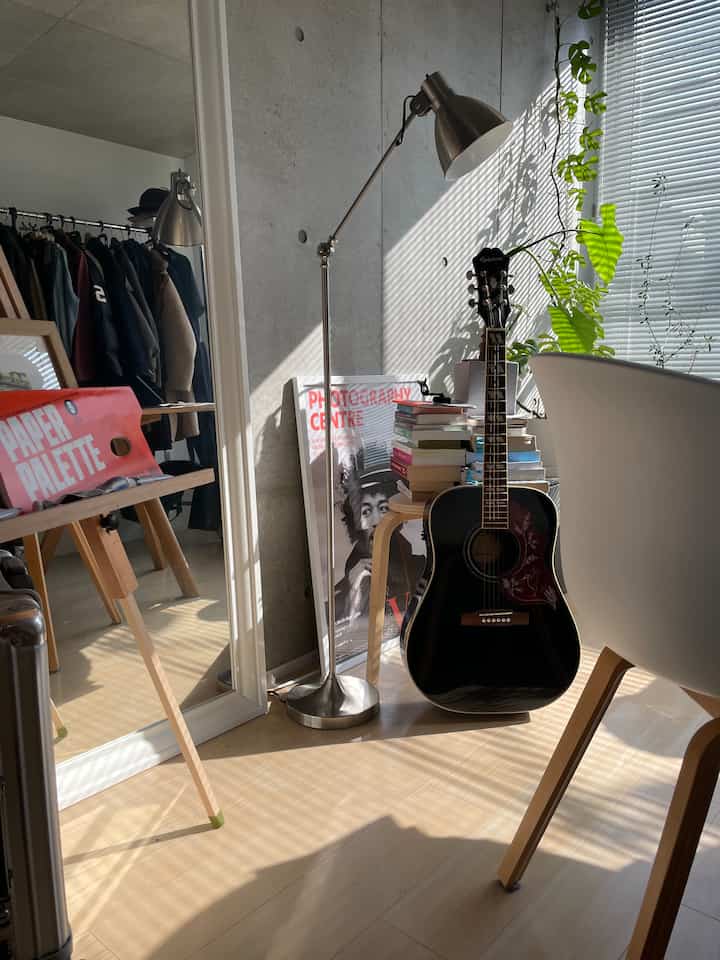 Modern compact room with concrete wall and wood floor, featuring stacked books on a stool and a black guitar in a bright cozy study space