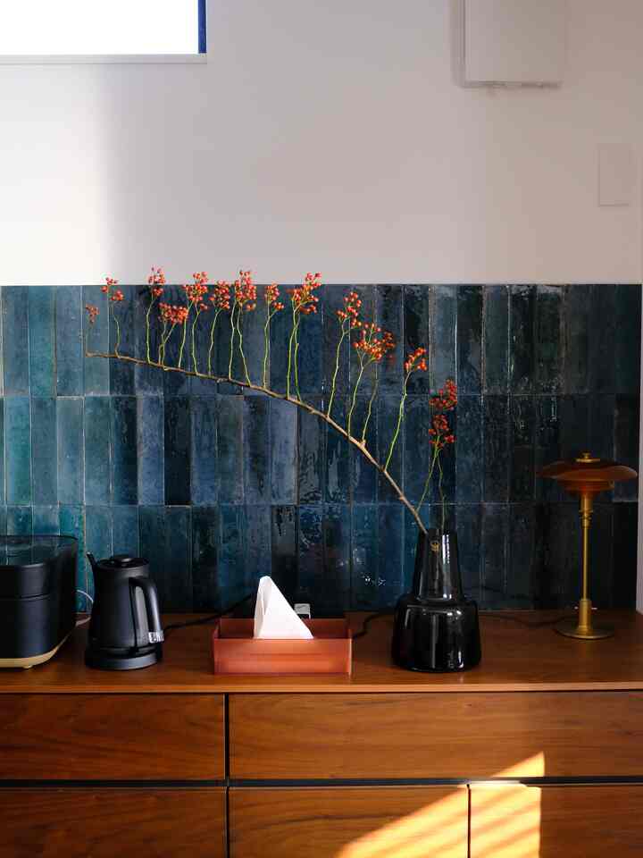 青いタイルの壁を背景に、木目調のキャビネットの上に黒い花瓶とキッチン用品が置かれた温かみのあるキッチン空間