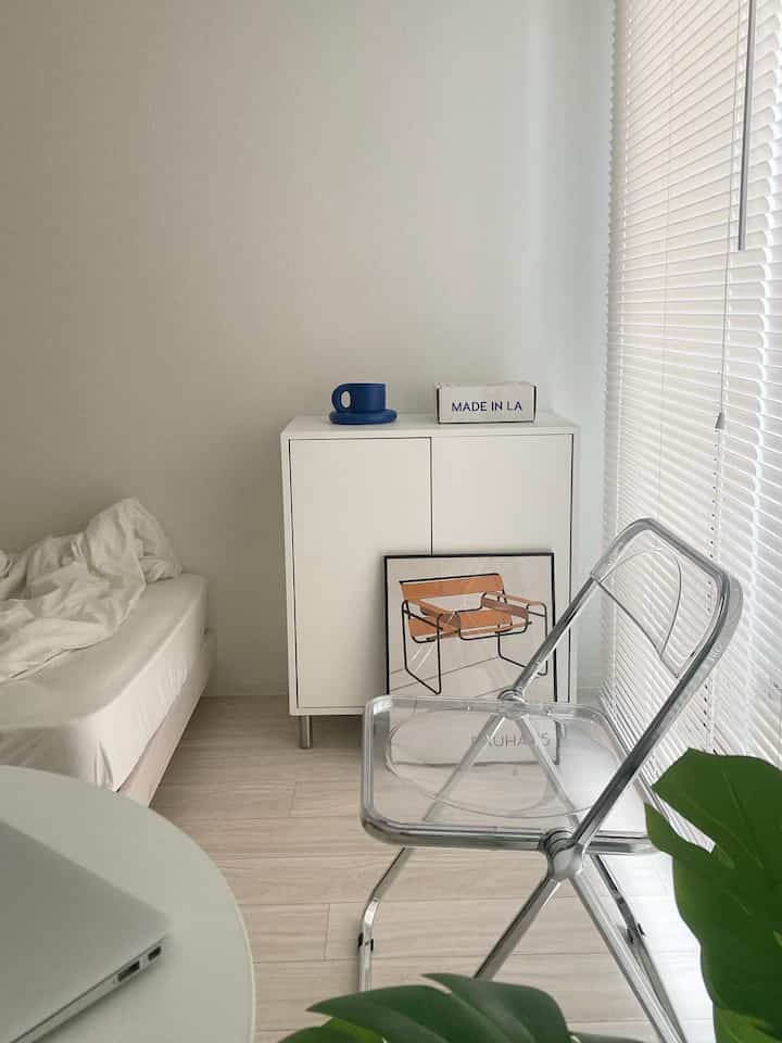 White and beige toned small bedroom featuring a transparent clear chair and a compact storage cabinet in a minimalist setting