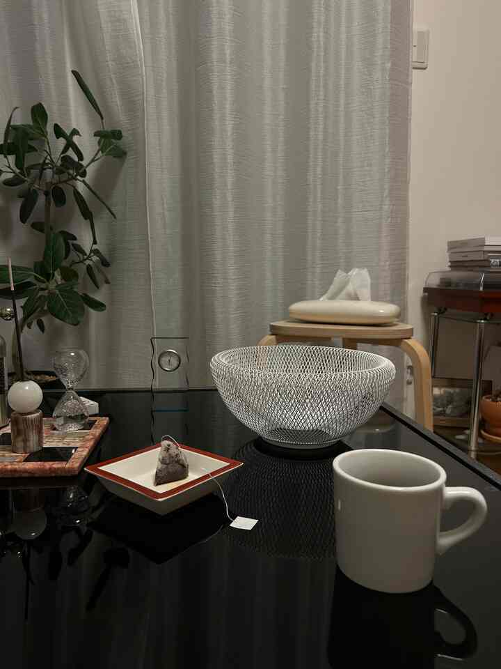 白いカーテンと植物があるリビング、中央に黒いガラスのコーヒーテーブルと白いメッシュのフルーツバスケットが置かれた雰囲気のある空間