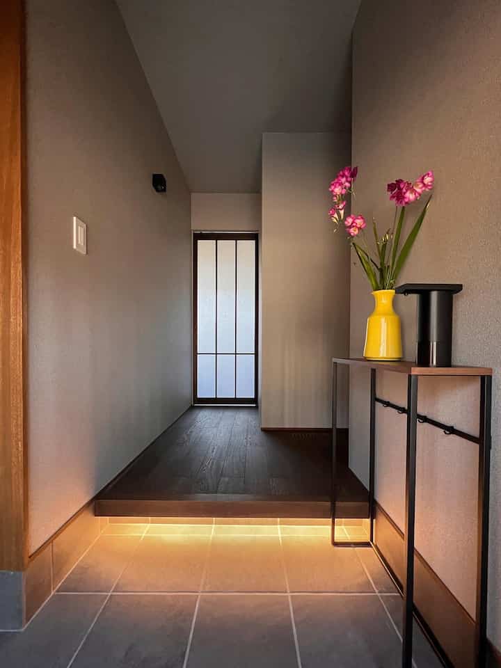 ブラウンの木製床とグレーの壁面がある狭いシンプルな玄関、コンソールテーブルに黄色の花瓶とオートディスペンサーが配置された空間