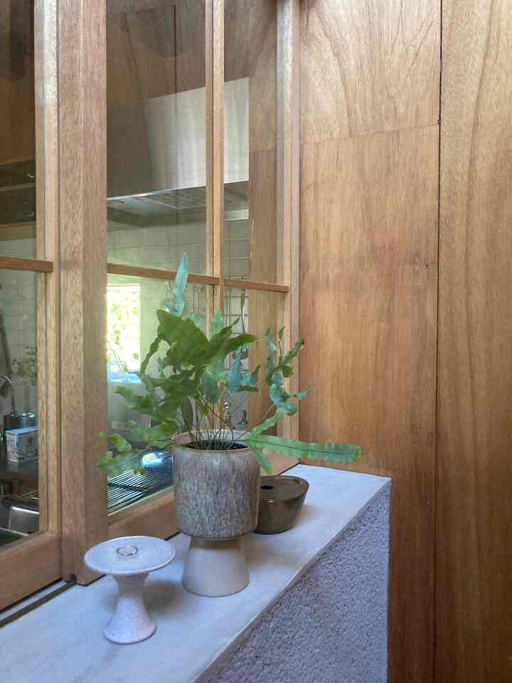 ナチュラルな木目調の壁と窓があるキッチン横の玄関空間、植物が置かれた小さなディスプレイ棚が見える