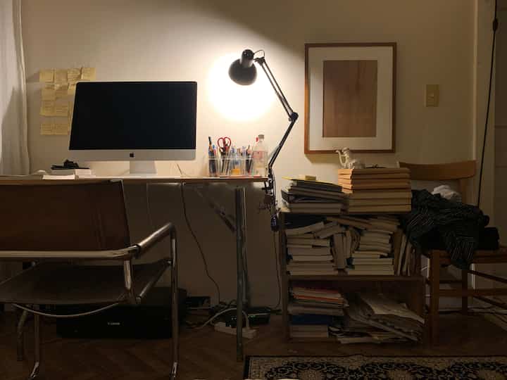 暖かみのあるブラウンとホワイトのトーンが特徴のワンルーム書斎、コンピューターデスクとランプ、本が整然と積まれたシンプルなホームオフィス