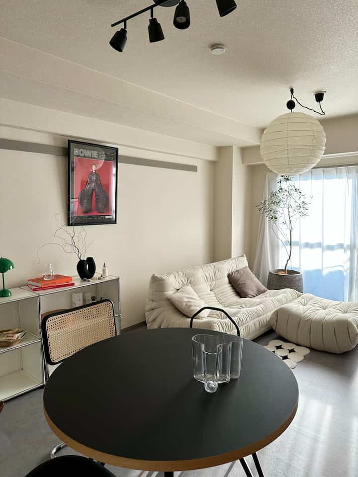 明るい白色の壁と窓辺にクリーム色のソファ、黒い丸テーブルの上に透明なガラス花瓶が置かれたミッドセンチュリーモダン風のリビング空間