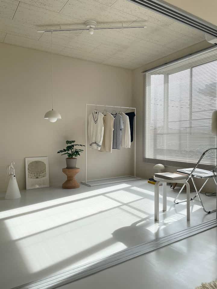 白を基調としたワンルーム、シンプルなアイアンハンガーラックとスツール、ブラインドが配置された雰囲気のあるインテリア