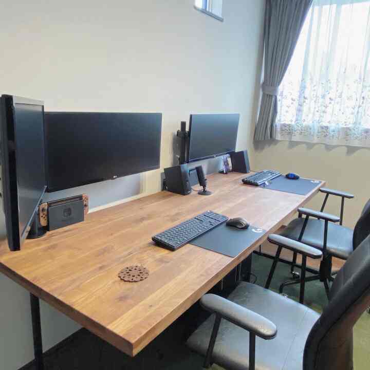 木目調のホームオフィス兼ゲームルーム、モニターアームに取り付けられた二つのモニターと黒いオフィスチェアが配置された清潔感のある空間