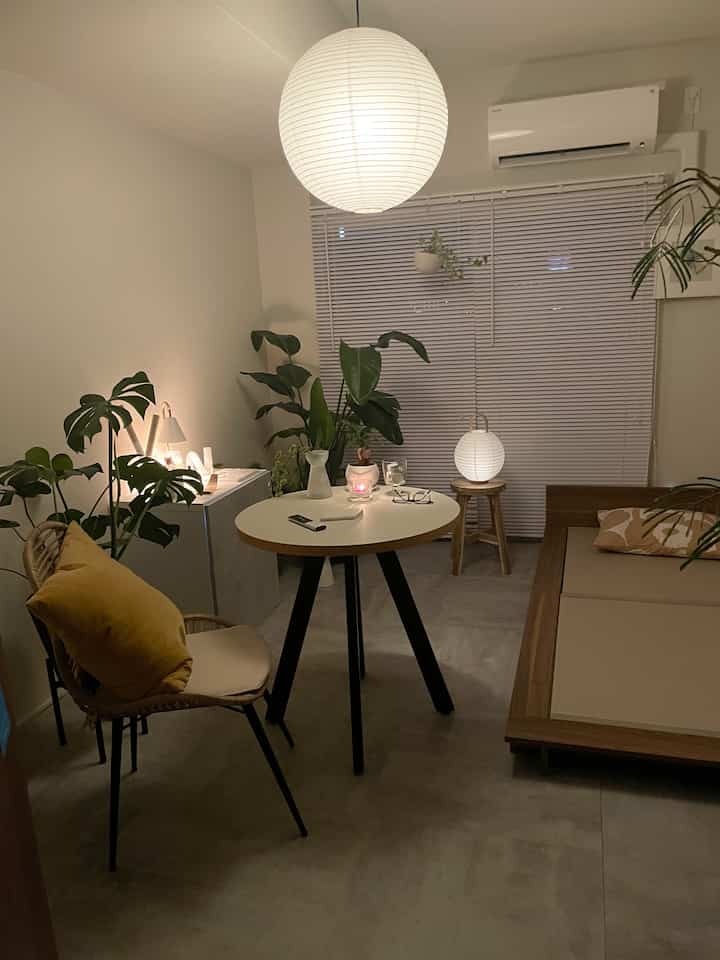 Small single household room with natural wood tones and green plants, featuring warm table lamp and candle light creating a cozy atmosphere