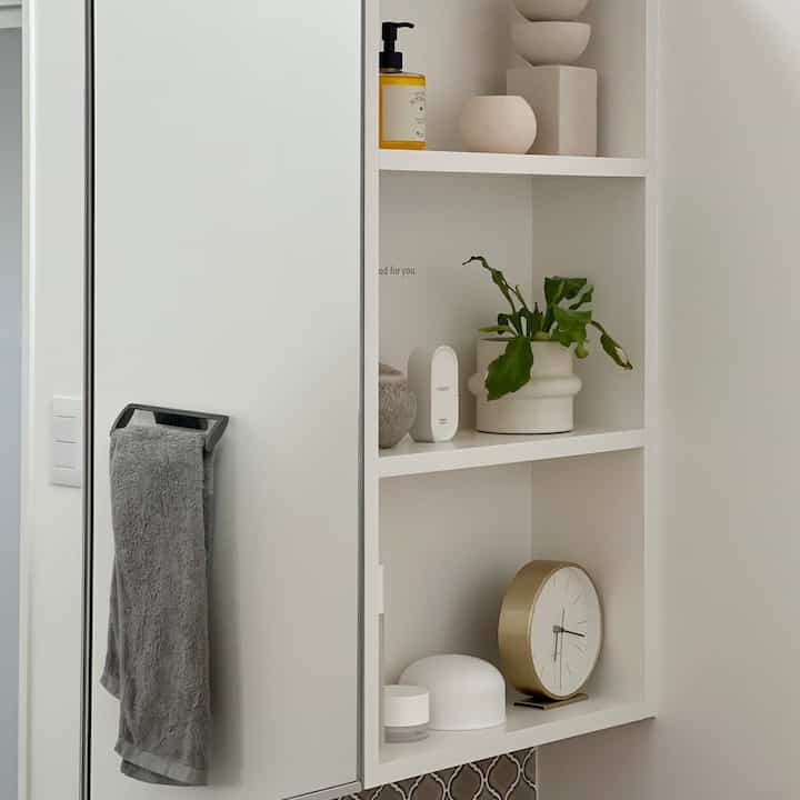 白とゴールドのトーンのパウダールームの棚、時計や植物、タオル掛けが配置された清潔感のある空間