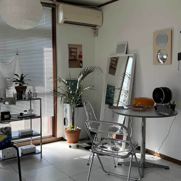 白を基調としたコンパクトなダイニングスペース、透明な椅子と大きな鏡、植物が配置された清潔感のあるモダンな空間