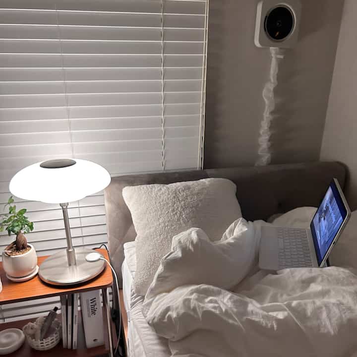 白を基調とした寝室、窓際にテーブルランプと植物が置かれたサイドテーブルと、ベッド上にノートパソコンがある雰囲気のある空間