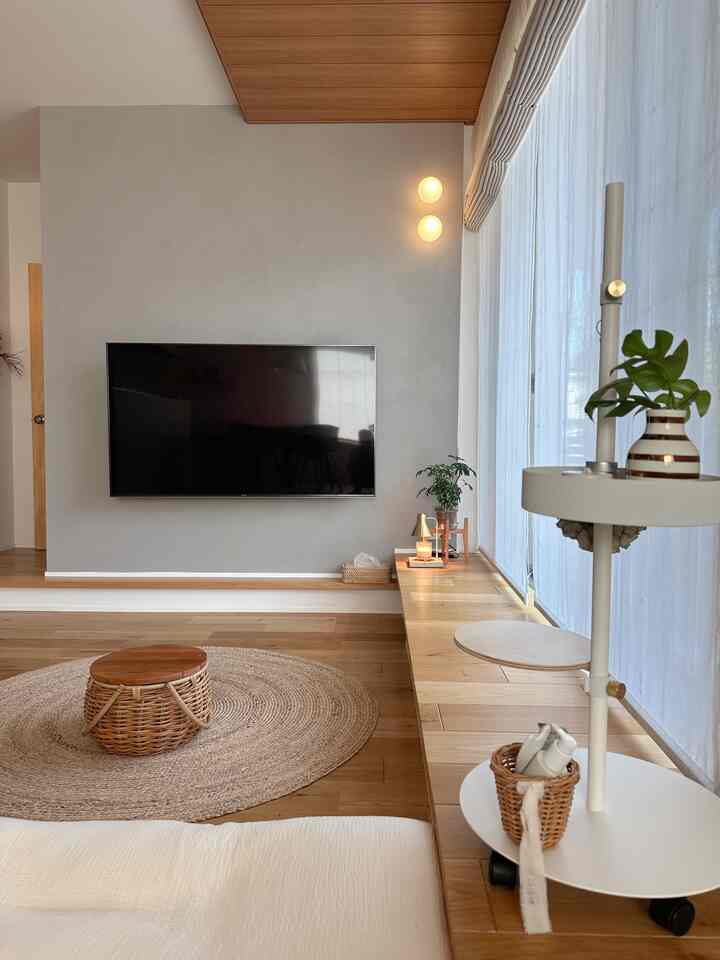 ナチュラルでウッドトーンのリビング、壁掛けテレビと長いベンチ、丸いラタンテーブルが配置されたシンプルな空間