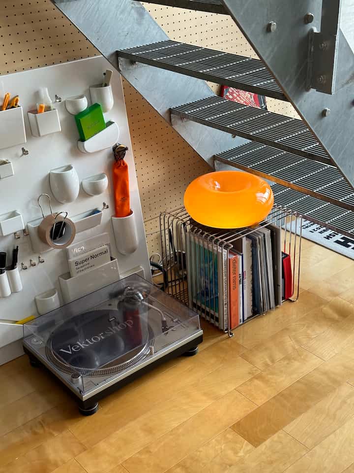 A compact space under metal stairs with wood-toned flooring featuring an orange donut-shaped stool, turntable, and bookshelf