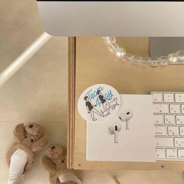 白とベージュのトーンのデスクの上に白いワイヤレスイヤホンの箱とキーボードがあり、床には犬の形をしたかわいいベージュ色のスリッパを履いた足が見える暖かな空間