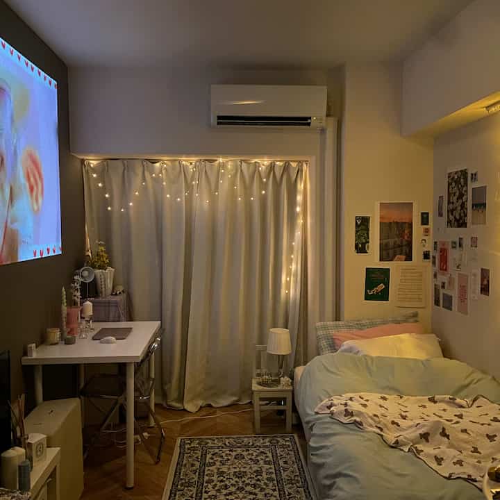 Cozy small studio apartment with warm lighting, featuring a bed, desk, and curtains decorated with string lights